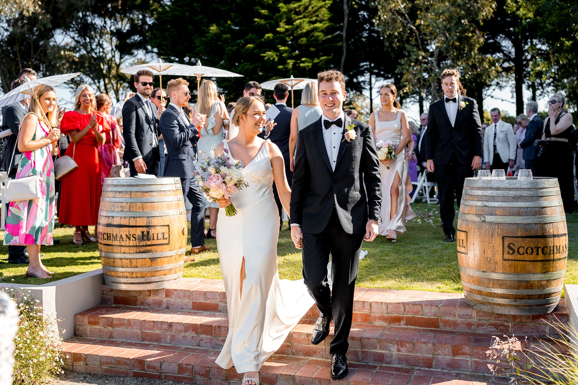 Bride and groom walking down steps after their ceremony at Scotchmans Hill winery, smiling as guests applaud with wine barrels in view.