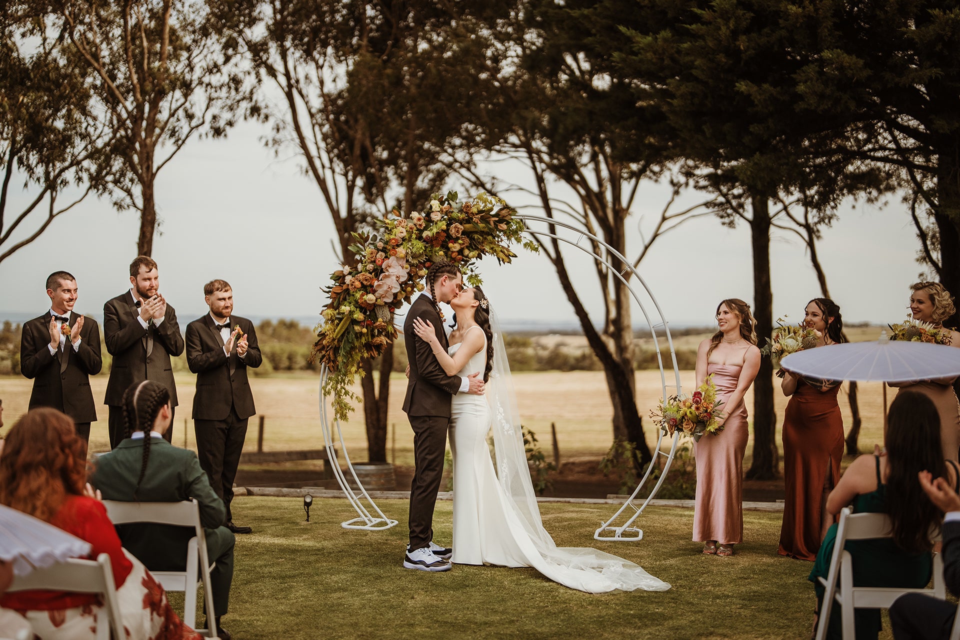 Couple sharing their first kiss under a floral arbour at Scotchmans Hill surrounded by their bridal party.