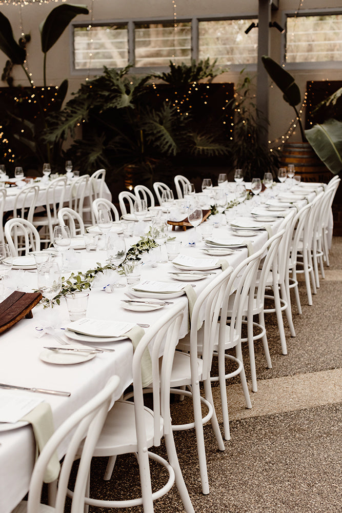 Long white tables with white bentwood chairs set for a wedding reception at Scotchmans Hill with greenery decor.