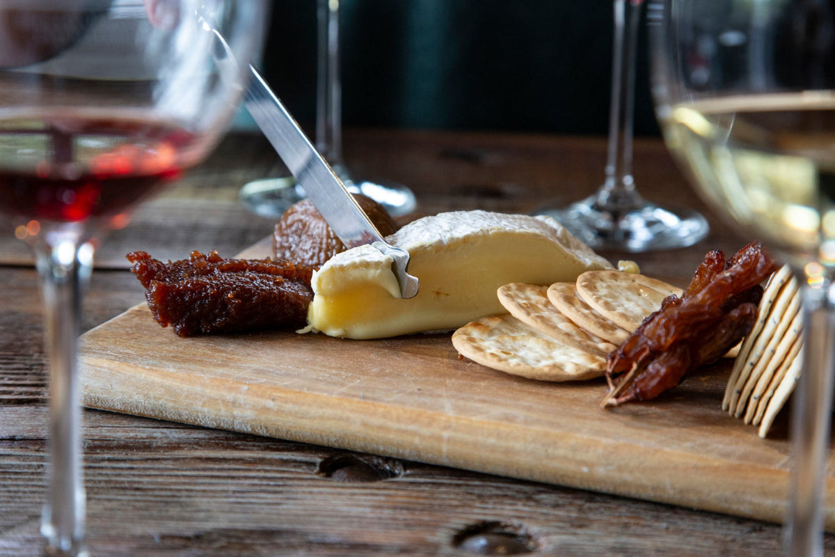 Wooden cutting board with cheese, crackers next to wine glasses.