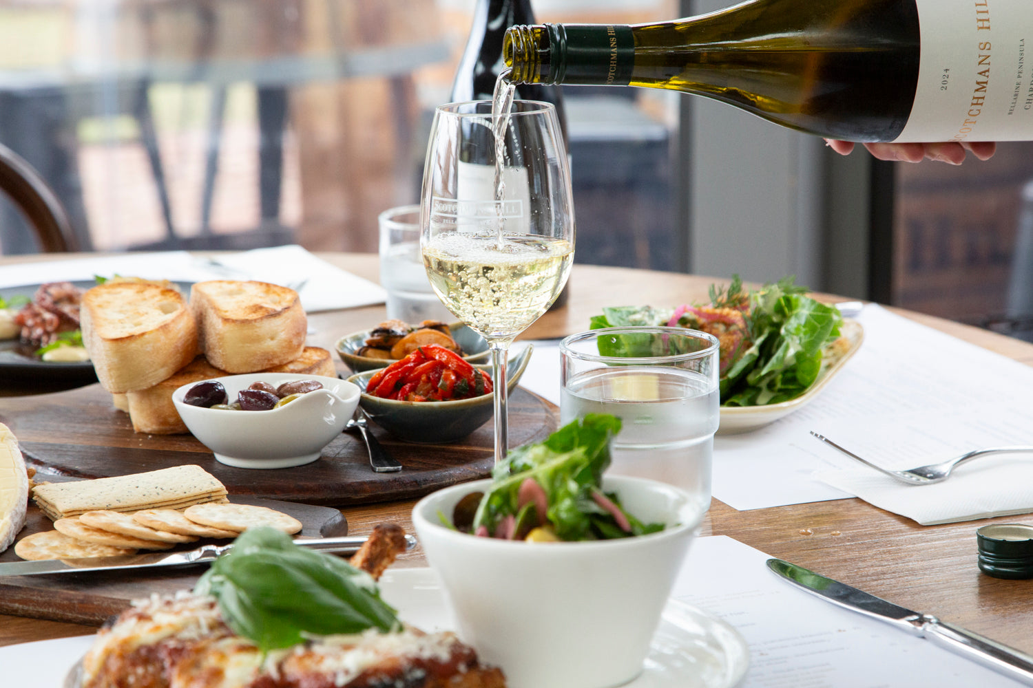 Person pouring white wine into a glass on a table with appetizers