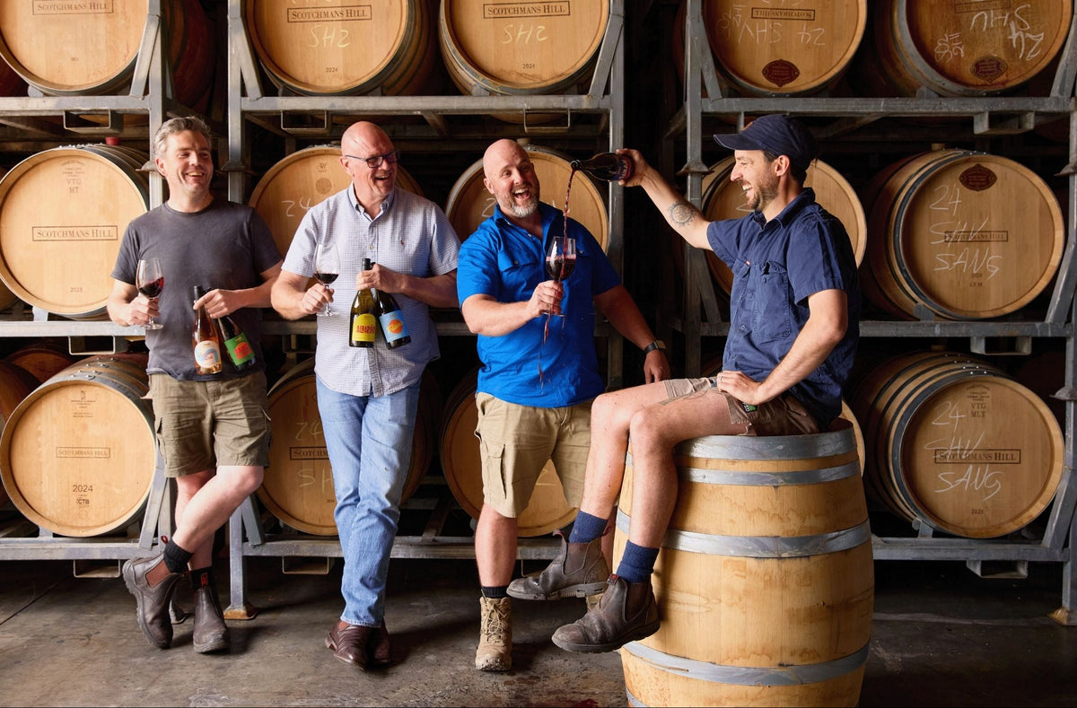 Scotchmans Hill winemakers standing in the barrel room holding Solé wines, laughing and pouring red wine with oak barrels stacked behind them.